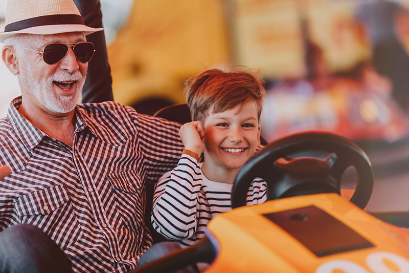 Mature, smiling man in sunglasses and fedora rides in bumper car with smiling grandson