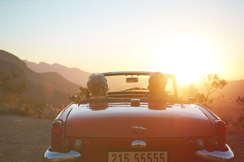 Mature laughing couple sitting in red convertible taking in sunset over mountain range